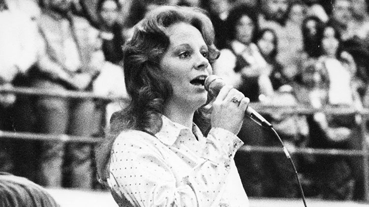 A black and white photo of Reba McEntire singing with the crowd standing up with their hands over their chests behind her.